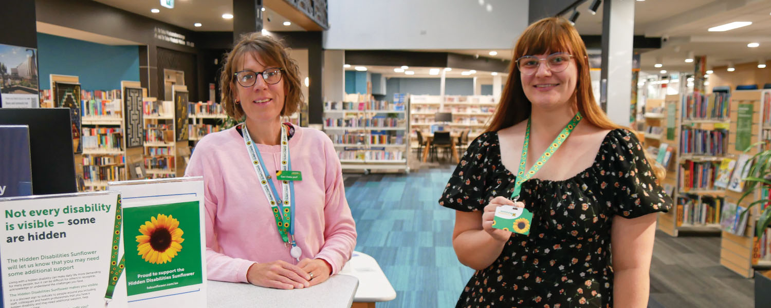 Two ladies wearing a sunflower lanyard standing next to a promotion stand, with library bookshelves in the background