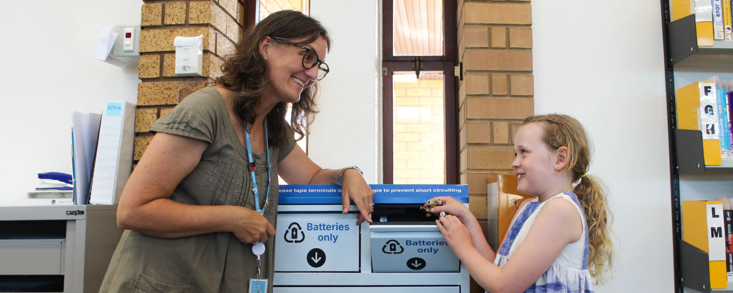 A smiling lady helping a child recycle batteries at the library